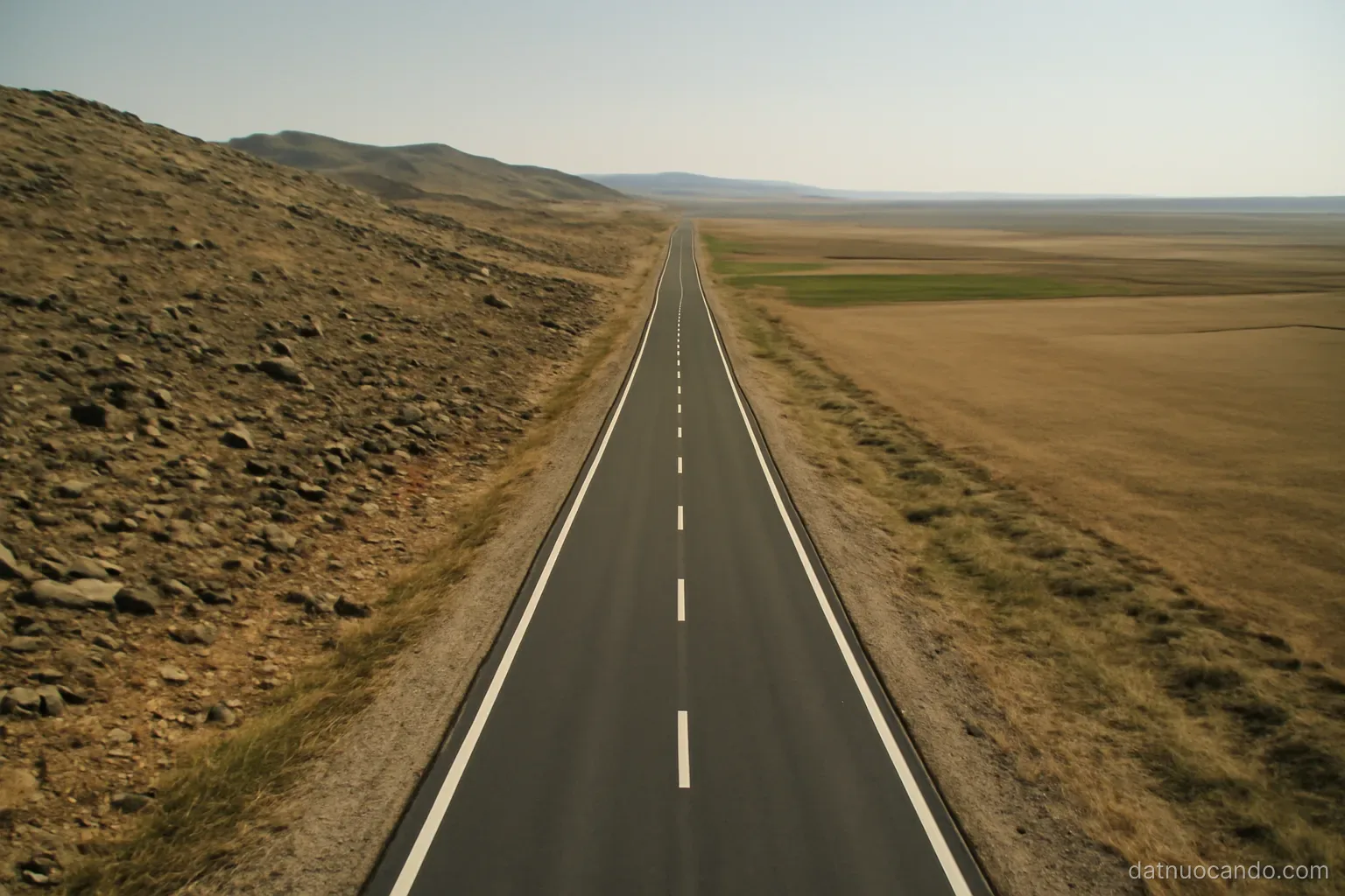 A long road cutting across three visibly different ground surfaces in one frame, rocky, hilly and flat, seen from a drone