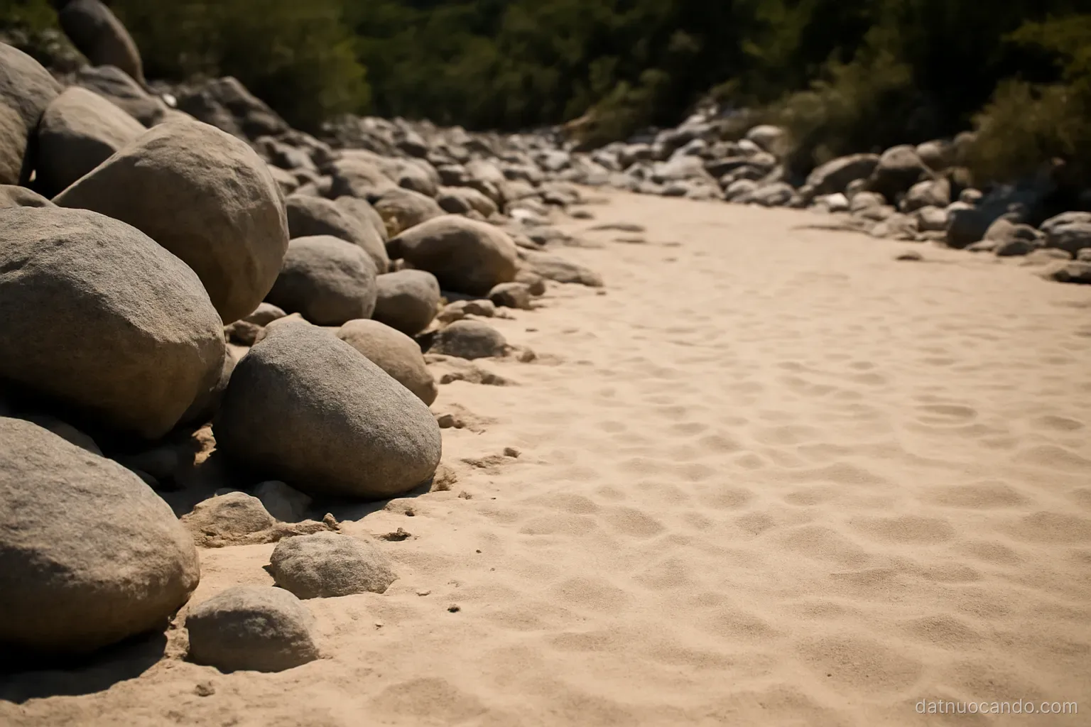 A dry riverbed where large boulders upstream give way to fine pale sand downstream, seen along its length at midday