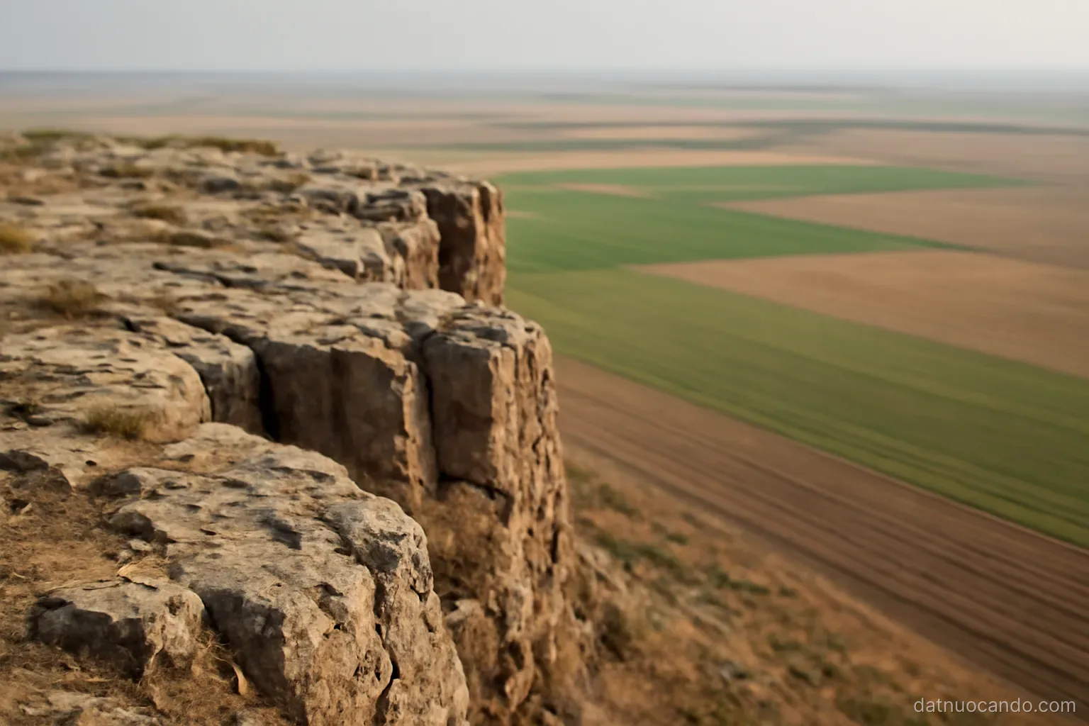 A rocky plateau edge meeting flat alluvial fields below, the boundary between them visible as a change in colour